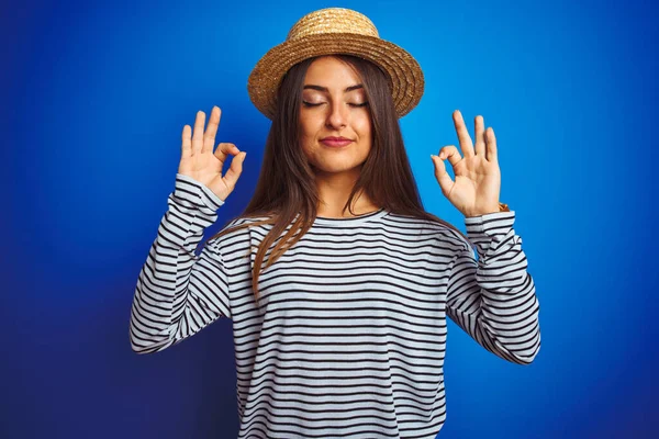 Young beautiful woman wearing navy striped t-shirt and hat over isolated blue background relax and smiling with eyes closed doing meditation gesture with fingers. Yoga concept.