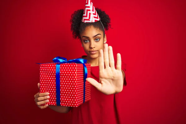 African american woman wearing funny hat holding birthday gift over isolated red background with open hand doing stop sign with serious and confident expression, defense gesture