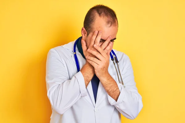 Young doctor man wearing coat and stethoscope standing over isolated yellow background with sad expression covering face with hands while crying. Depression concept.