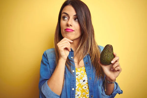 Giovane Bella Donna Mangiare Avocado Sano Sfondo Giallo Volto Serio — Foto Stock