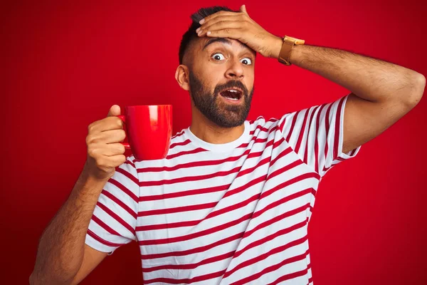 Young indian man wearing striped t-shirt drinking cup of coffee over isolated red background stressed with hand on head, shocked with shame and surprise face, angry and frustrated. Fear and upset for mistake.