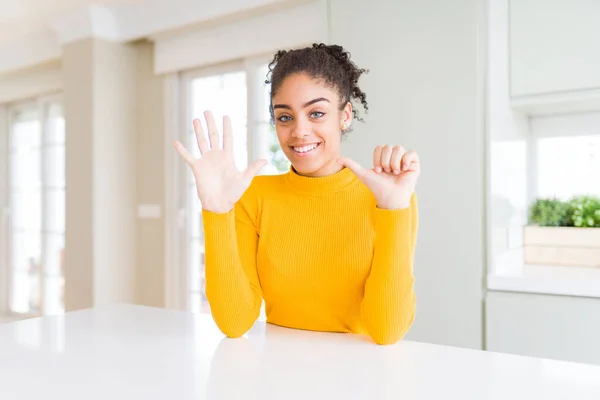Beautiful african american woman with afro hair wearing a casual yellow sweater showing and pointing up with fingers number six while smiling confident and happy.