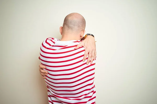 Young bald man with beard wearing casual striped red t-shirt over white isolated background Hugging oneself happy and positive from backwards. Self love and self care