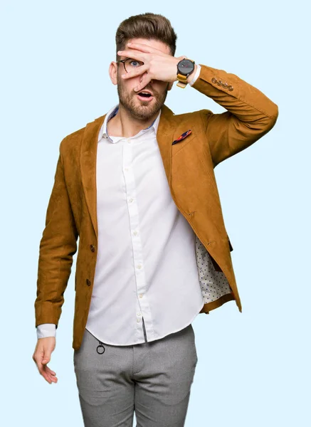 Young handsome business man wearing glasses peeking in shock covering face and eyes with hand, looking through fingers with embarrassed expression.