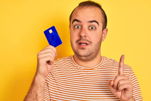 年轻男子拿着蓝色的信用卡 站在孤立的黄色背景上 惊讶地发现一个想法或问题 满脸喜形于色 指手画脚 位居第一 — 图库照片