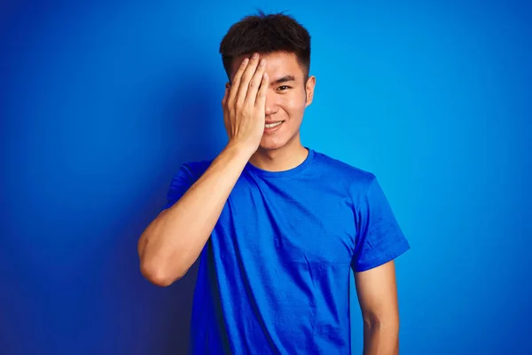 Tシャツを着た若いアジア系中国人男性が 片方の目を手で覆い 顔に自信を持って笑顔と驚きの感情を覆う孤立した青い背景の上に立って — ストック写真
