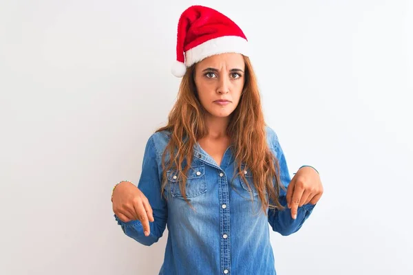 若い美しい赤毛の女性は孤立した背景の上にクリスマスの帽子をかぶっている指で方向を示す 悲しいと動揺を下に向けて 不幸なと落ち込んで — ストック写真