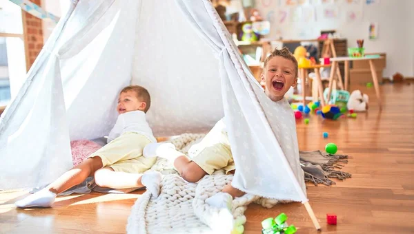 Adorable blonde twins playing inside tipi around lots of toys at kindergarten