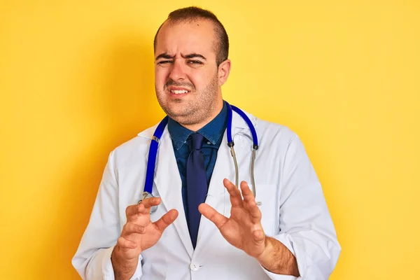 Young doctor man wearing coat and stethoscope standing over isolated yellow background disgusted expression, displeased and fearful doing disgust face because aversion reaction. With hands raised. Annoying concept.