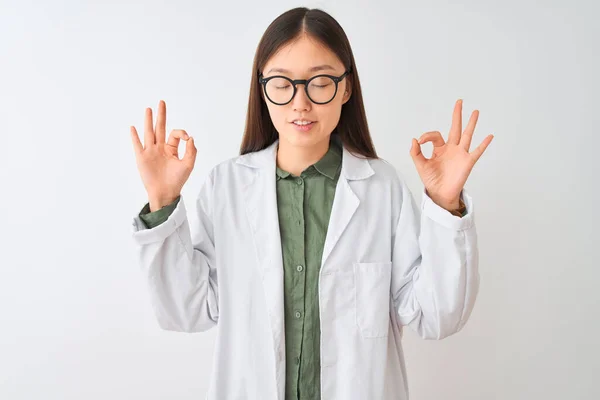 Young chinese scientist woman wearing coat and glasses over isolated white background relax and smiling with eyes closed doing meditation gesture with fingers. Yoga concept.