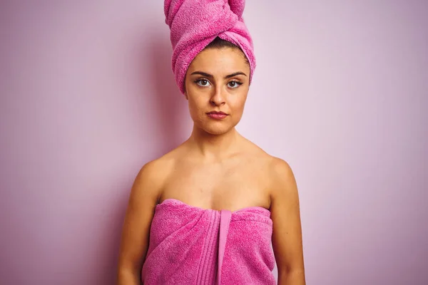 Young beautiful woman wearing towel after shower over isolated pink background with serious expression on face. Simple and natural looking at the camera.