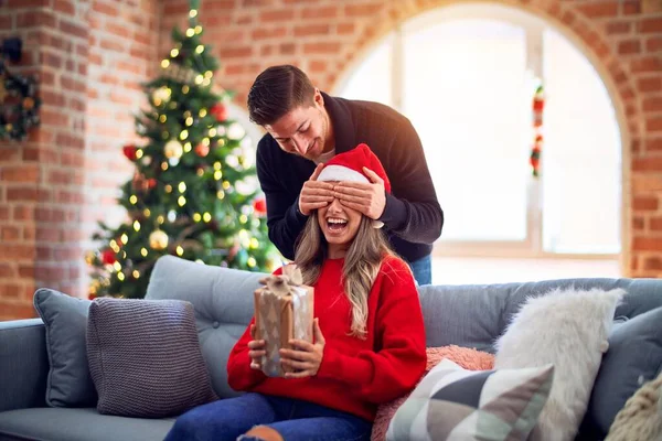 年轻漂亮的夫妇面带微笑 既快乐又自信 男人蒙住女人的眼睛 在家里的圣诞树旁 用礼物来抚慰她 — 图库照片