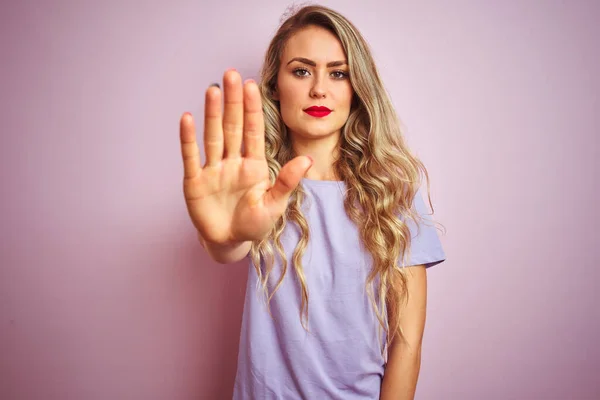 Young beautiful woman wearing purple t-shirt standing over pink isolated background doing stop sing with palm of the hand. Warning expression with negative and serious gesture on the face.