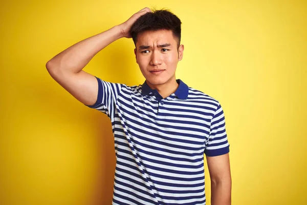 Young asian chinese man wearing striped polo standing over isolated yellow background confuse and wonder about question. Uncertain with doubt, thinking with hand on head. Pensive concept.