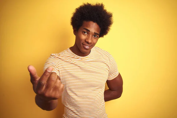 American man with afro hair wearing striped t-shirt standing over isolated yellow background Beckoning come here gesture with hand inviting welcoming happy and smiling