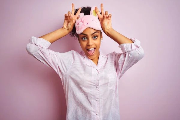 年轻的非洲裔美国女人 戴着睡衣和面罩 披着孤立的粉色背景 头戴滑稽而疯狂的手指 就像兔子的耳朵 开心地微笑着 — 图库照片