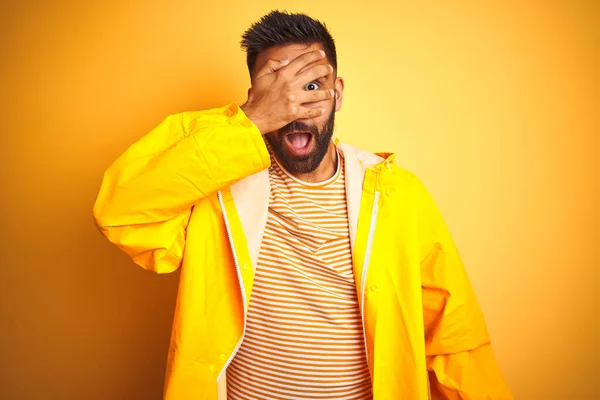 Young indian man wearing raincoat standing over isolated yellow background peeking in shock covering face and eyes with hand, looking through fingers with embarrassed expression.