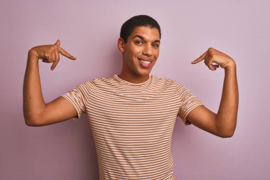 Genç, çizgili tişört giyen yakışıklı Arap adam izole edilmiş pembe arka planda duruyor. Yüzünde bir gülümsemeyle, gururlu ve mutlu bir şekilde kendini gösteriyor..