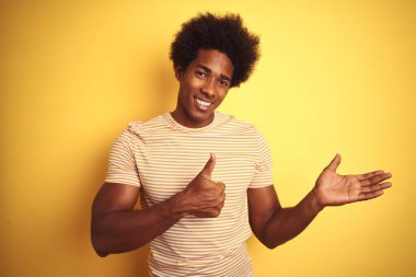 Afro saçlı, çizgili tişörtlü, izole edilmiş sarı arka planda duran, avuçlarını gösteren ve baş parmaklarıyla hareket eden mutlu ve neşeli gülümseyen bir Amerikalı.