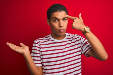İzole edilmiş kırmızı arkaplan üzerine çizgili tişört giyen genç yakışıklı Arap adam avuç içinin açık olmasından rahatsız ve kafası karışmış bir şekilde kopya alanı gösteriyor ve alnını işaret ediyor. Bunu bir düşün..