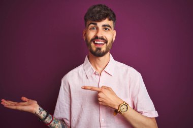 Yalıtılmış mor arka plan üzerinde duran pembe tişörtlü genç adam kameraya hayret ediyor ve gülümsüyor..