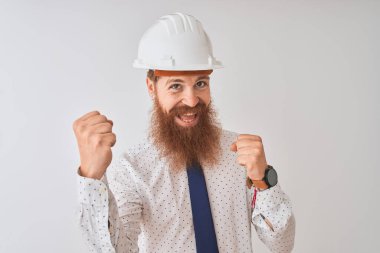 İzole edilmiş beyaz arka plan üzerinde güvenlik kaskı takan genç kızıl saçlı mimar çok mutlu ve heyecanlı bir şekilde kollarını kaldırıp, gülümseyerek ve başarı için çığlık atarak zafer hareketi yapıyor. Kutlama konsepti.