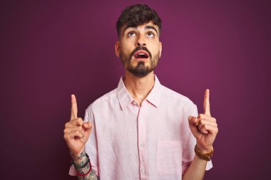 Yalıtılmış mor arka plan üzerinde duran pembe tişörtlü genç adam şaşırmış ve şaşırmıştı. Parmaklarıyla işaret ediyor ve kollarını kaldırıyordu..