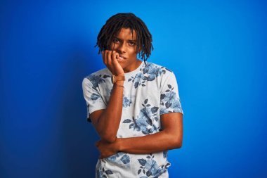 Rasta desenli tişörtlü Afro-Amerikan bir adam izole edilmiş mavi arka plan üzerinde gergin ve gergin görünüyor ve elleri ağızdan tırnak yiyor. Endişe sorunu.