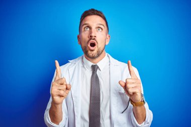 İzole edilmiş arka plan üzerinde beyaz profesyonel ceket giyen yakışıklı doktor şaşırmış ve şaşırmış bir şekilde yukarı baktı ve parmaklarıyla işaret etti ve kollarını kaldırdı..