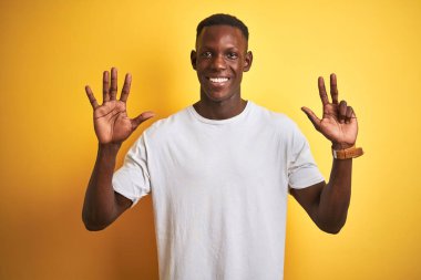 Beyaz t-shirt giyen genç Afro-Amerikalı adam izole edilmiş sarı arka plan üzerinde duruyor ve 8 numarayı işaret ederek kendinden emin ve mutlu gülümsüyor..