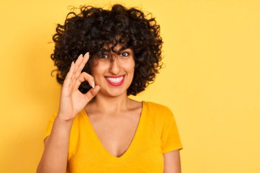 Sarı arka planda duran kıvırcık saçlı, kıvırcık saçlı genç Arap kadın. Tamam işareti yapıyor, mükemmel bir sembol.