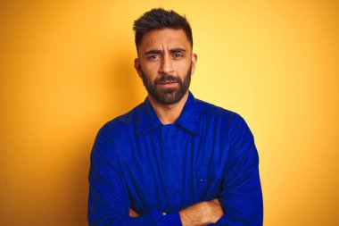 İzole edilmiş sarı arka plan şüphecisi ve gergin, yüzünde çapraz kollu tasvip etmeyen ifade üzerine üniforma giyen yakışıklı Hintli işçi. Negatif kişi.