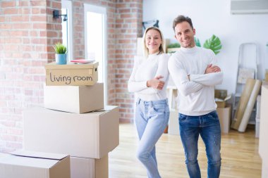 Karton kutuların etrafında durmuş kollarını kavuşturup kameraya bakan mutlu yüzlü genç bir çift. Pozitif kişi.