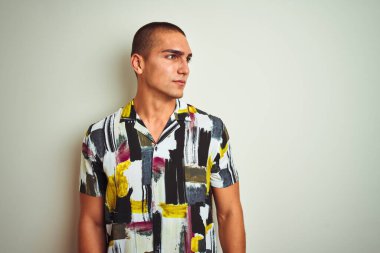 Beyaz tenli arka planda yaz tişörtü giyen yakışıklı bir adam yüzünde gülümseme, doğal bir ifade ile yan yana bakıyor. Kendinden emin gülen.