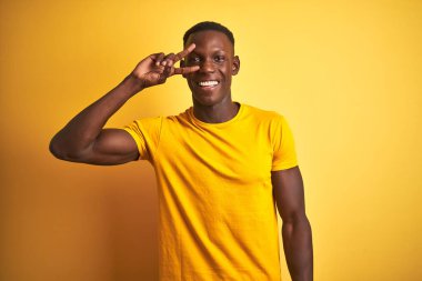 İzole sarı arka plan üzerinde ayakta rahat t-shirt giyen genç Afrikalı Amerikalı adam yüz üzerinde parmaklarla barış sembolü yapıyor, neşeli gösteren zafer gülümseyen