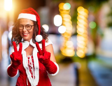 Orta yaşlı kadın Noel Baba kostümü giyip kameraya bakıp zafer işareti yapıyor. İki numara..