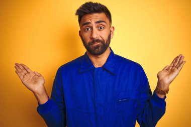 Sarı arka plan üzerinde üniforma giyen yakışıklı Hintli işçi elleri ve kolları havada, kafası karışık bir ifade takınıyor. Şüphe kavramı.