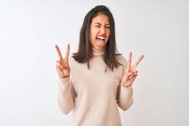 Balıkçı yaka kazak giyen güzel Çinli kadın izole edilmiş beyaz arka planda dikiliyor ve iki elini de zafer işareti yaparken gösteren bir dille gülümsüyor. İki numara..