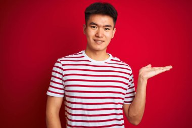 Asyalı genç bir Çinli, çizgili tişört giyiyor. Kırmızı arka planda duruyor. Gülümsüyor, avucunun içi gibi kameraya bakıyor..