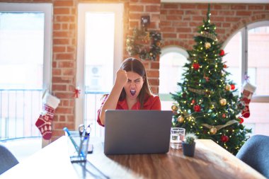Güzel bir kadın evde dizüstü bilgisayarla oturup Noel ağacının etrafında kızgın ve kızgın bir şekilde çalışıyor öfkeyle bağırırken yumruğunu kaldırıyor. Öfke ve agresif kavram.
