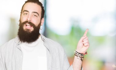 Uzun saçlı, sakallı ve küpeli genç adam yüzünde kocaman bir gülümseme ile, kameraya bakarak yan el ve parmak ile işaret.