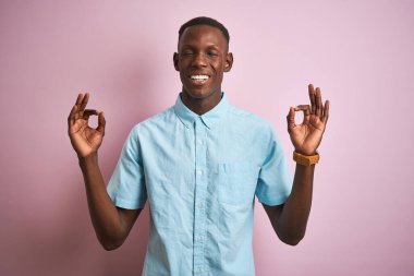 Mavi gömlekli Afro-Amerikalı bir adam pembe arka planda dikiliyor rahatlıyor ve parmaklarıyla meditasyon hareketi yaparken gözleri kapalı gülümsüyor. Yoga konsepti.