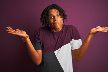 Rastgele tişörtler giyen Afro bir adam izole edilmiş mor arka plan üzerinde duruyor. Kolları ve elleri havada, kafası karışmış bir ifade. Şüphe kavramı.