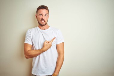 İzole edilmiş arka plan üzerinde gündelik beyaz tişört giyen genç yakışıklı bir adam endişeli ve gergin bir kenara işaret ediyor.