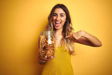 Sarı arka planda bir kavanoz kurabiyeyle gezen genç ve güzel bir kadın kendini gösteren sürpriz bir yüzle.