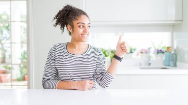 Afro saçlı, sıradan çizgili süveter giyen, gururlu, gülümseyen ve baş parmak hareketi yapan güzel Afrikalı Amerikalı kadın.