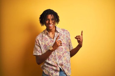 Sarı arka planda çiçek desenli gömlekli Afro adam gülümsüyor ve kameraya bakıyor. İki eliyle ve parmaklarıyla yan tarafa bakıyor..