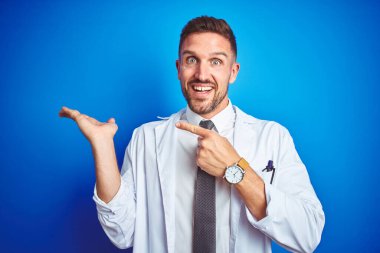 İzole arka plan üzerinde beyaz profesyonel ceket giyen genç yakışıklı doktor adam şaşırttı ve el ile sunarken kameraya gülümseyerek ve parmak ile işaret.