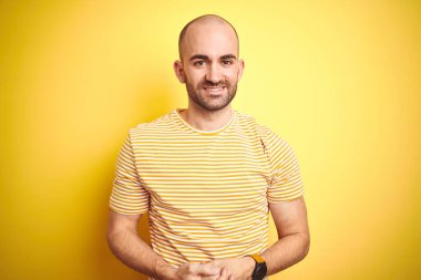 Sarı izole arka plan üzerine sıradan çizgili tişört giyen sakallı genç ve kel bir adam ellerini kavuşturup gülümsüyor. Başarı ve iyimser