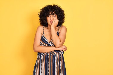 Sarı arka planda çizgili elbise giyen kıvırcık saçlı Arap bir kadın. Ağzı tırnak ısıran elleri yüzünden gergin ve stresli görünüyor. Endişe sorunu.